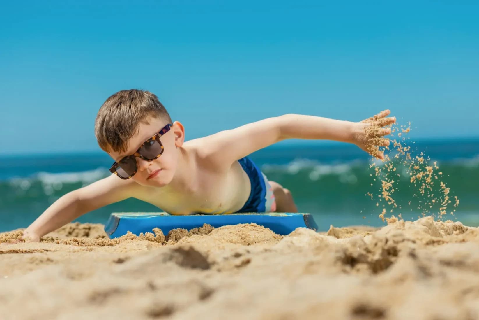 a young boy with sunglasses laying down on a body board on a beach